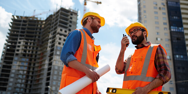 “Bauunternehmenmitarbeiter” Zwei Baustellen Mitarbeiter in Warnwesten und Bauhelmen sprechen über Funk mit dem Bauleiter auf einer Baustelle