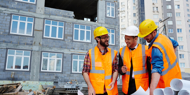 “Baustellenversammlung” Drei Bauarbeiter vor einer Hochhaus Baustelle in oragen Warnwesten zusammen mit dem Baulleiter mit einem weißen Bauhelm