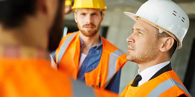 “Baubesprechung-der-Kubau-GmbH” Drei Bauleiter in Warnwesten und mit Baustellenhelm während einer Baubesprechung auf einer Baustelle in Kiel