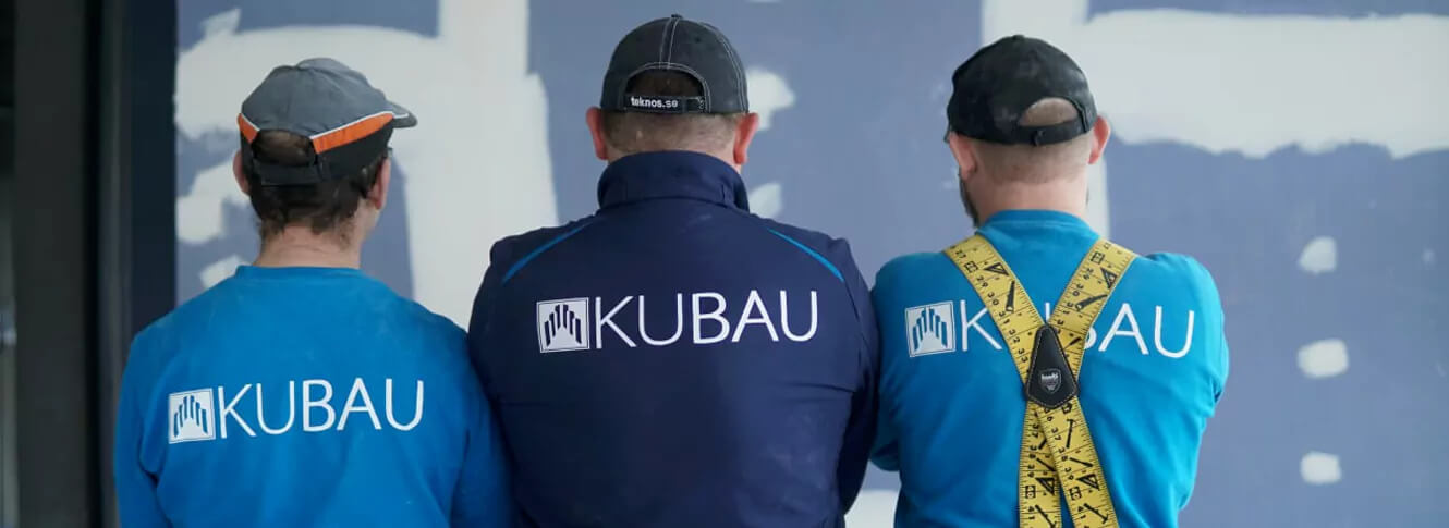 Drei Trockenbaumonteure sitzen mit dem Rücken vor einer Trockenbauwand mit dem Firmenschriftzug KUBAU