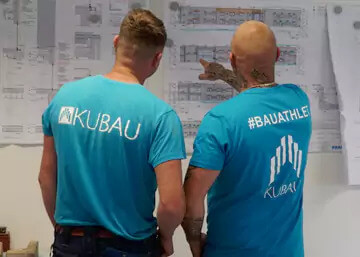Zwei Bauleiter mit Kubau Firmen Tshirt vor einem Baustellenplan an einer Trockenbauwand